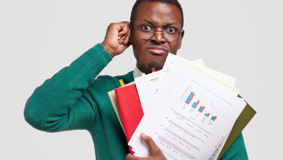 Dissatisfied black man prepares accounting report, holds papers with inforgraphic, wears spectacles for good vision, scratches head in bewilderment, isolated over white background. Income analysis