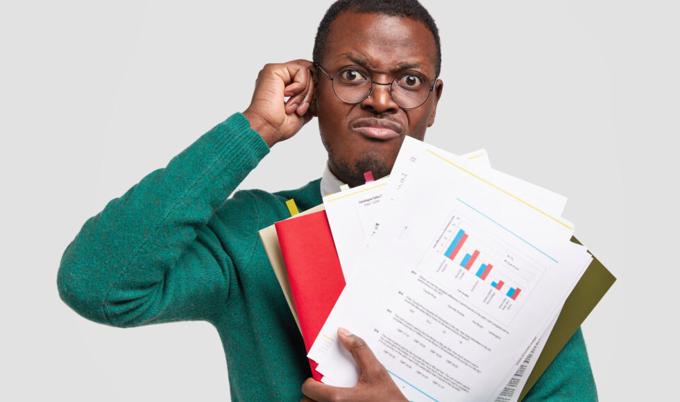 Dissatisfied black man prepares accounting report, holds papers with inforgraphic, wears spectacles for good vision, scratches head in bewilderment, isolated over white background. Income analysis Dissatisfied black man prepares accounting report, holds papers with inforgraphic, wears spectacles for good vision, scratches head in bewilderment, isolated over white background. Income analysis